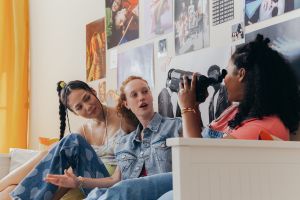 zwei junge Frauen werden von einer dritten Frau mit einer Kamera gefilmt und schauen in die Kamera © pexels-cottonbro